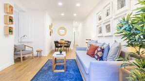 Ein modernes Wohnzimmer mit blauem Teppich, einem grauen Sofa mit bunten Kissen, einem Holztisch, einem runden Spiegel und einem Essbereich mit Tisch und Stühlen.