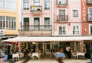 Vista da rua com um edifício rosa identificado como “OCCANDA” e uma área externa com assentos, mesas e cadeiras sob um toldo branco.