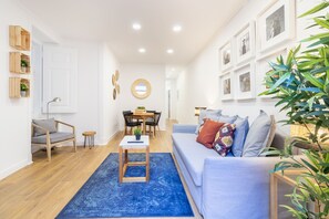 Uma sala moderna com tapete azul, sofá cinza com almofadas coloridas, mesa de centro de madeira, espelho redondo e um espaço de jantar com mesa e cadeiras.