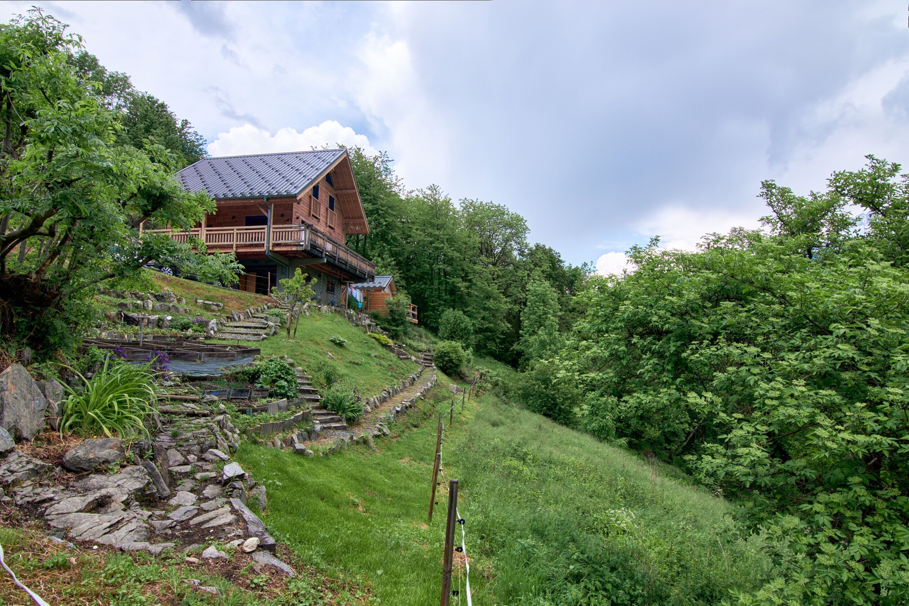 A wooden cabin with a balcony, surrounded by lush greenery and a small stream.