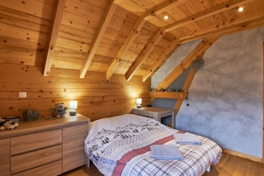 A wooden cabin bedroom with a sloped ceiling, a bed with patterned bedding, bedside tables with lamps, and a small shelf with decorative items. - Chalet "La Traverse" with Mountain View, Whirlpool & Wi-Fi (Allemond)