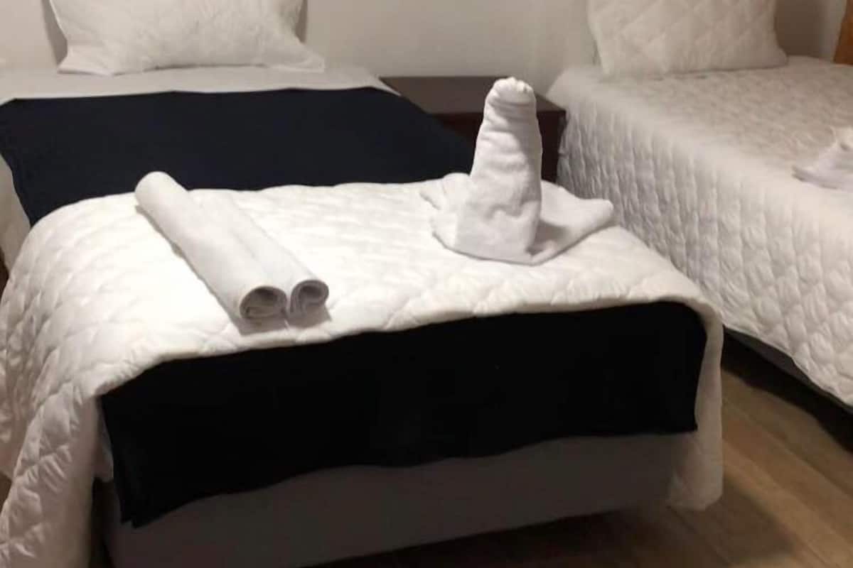 Habitación de hotel con dos camas, cada una con ropa de cama blanca y cabeceros oscuros. Se aprecia una toalla enrollada y un paño blanco en la cama del primer plano.