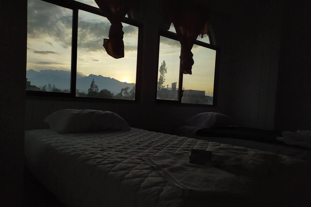 Una habitación tenuemente iluminada con una cama, dos ventanas y vistas a montañas y árboles.