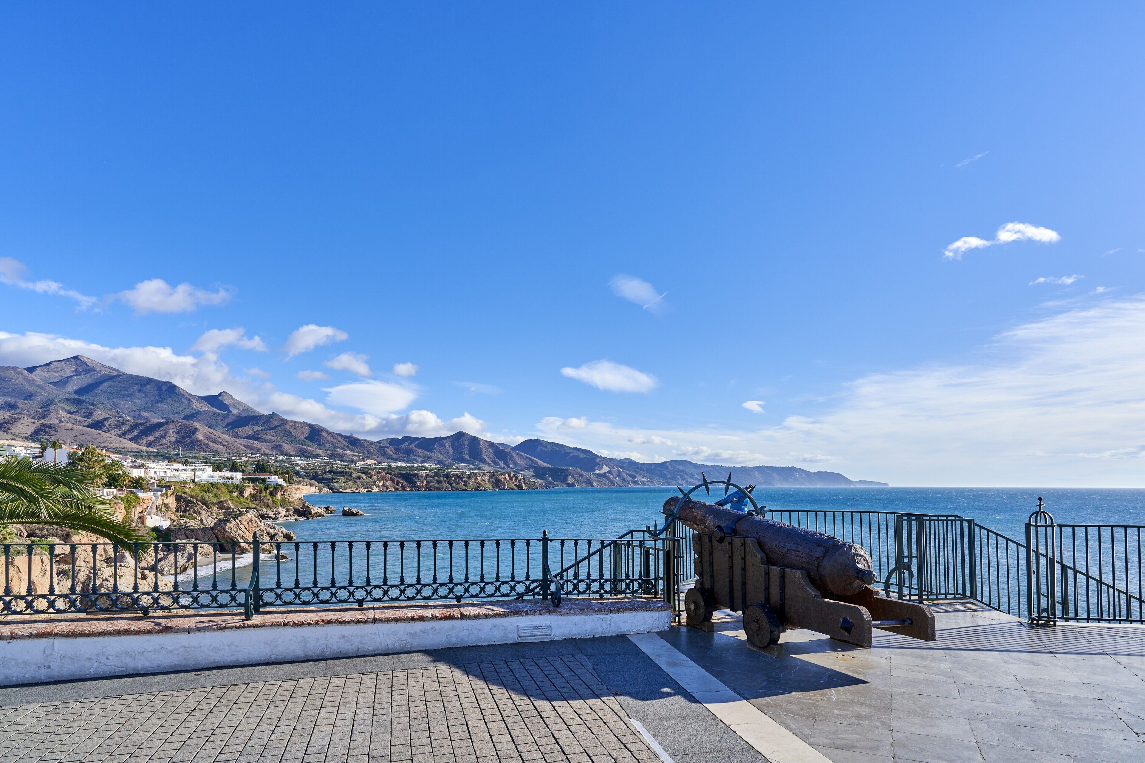 A coastal view with a cannon, a paved walkway, and a railing overlooking the sea and mountains.