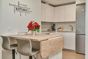 A modern kitchen with white cabinets, a wooden table, two chairs, a potted plant, and a stainless steel refrigerator.