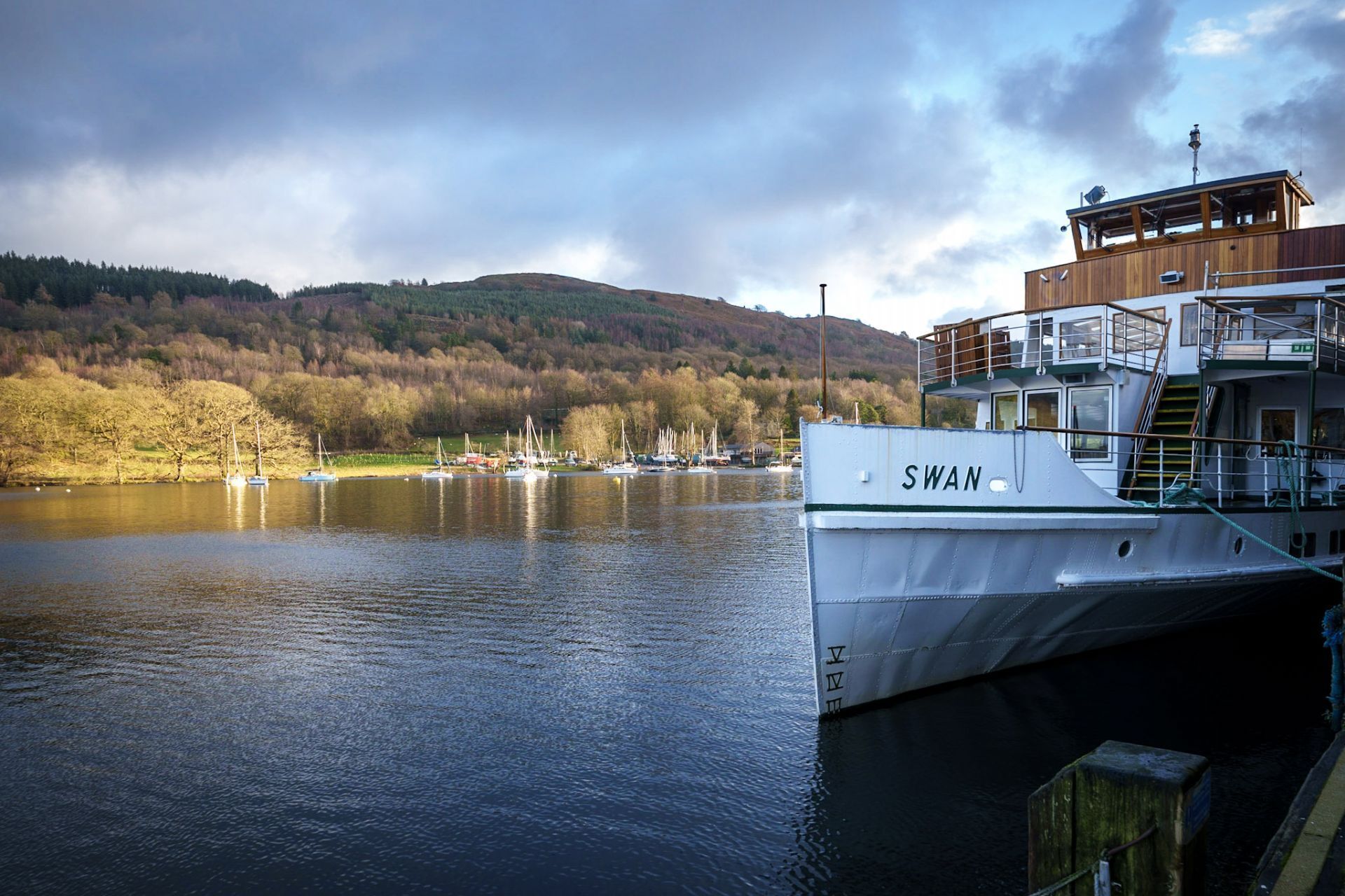 A boat named 'SWAN' docked at a pier with a view of a forested hillside in the background.
