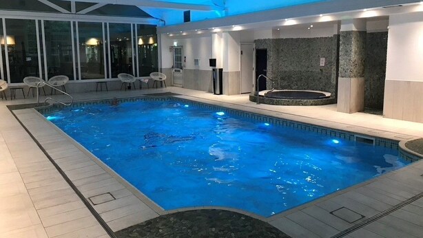 En indendørs swimmingpool med krystalklart blåt vand, omgivet af hvide vægge og et loft med blå belysning. Der er liggestole og et område med jacuzzi.