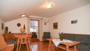 Ein gemütliches Wohnzimmer mit einem Holzesstisch, einer Couch und einem Sessel. Ein Fenster mit Vorhängen, einer Wandleuchte und einem Bilderrahmen sind ebenfalls zu sehen.