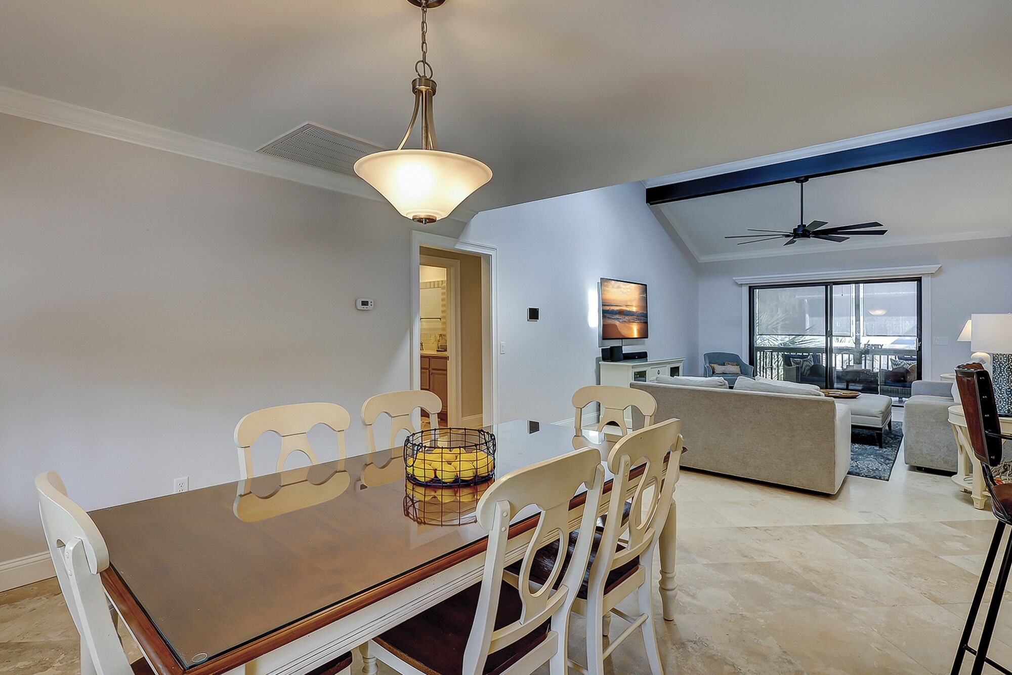 A modern living room with a large window, a dining area with a wooden table and white chairs, and a ceiling fan.