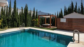 A swimming pool with a wooden deck and a swing chair, surrounded by tall trees and a mountain in the background. - RDR254 - Stunning 1 bedroom Apt. Golden Banús! (Marbella)