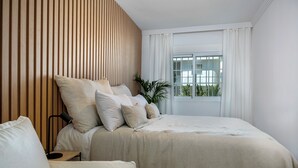 A bedroom with a large bed, a wooden slat wall, a window with curtains, and a potted plant. - RDR254 - Stunning 1 bedroom Apt. Golden Banús! (Marbella)