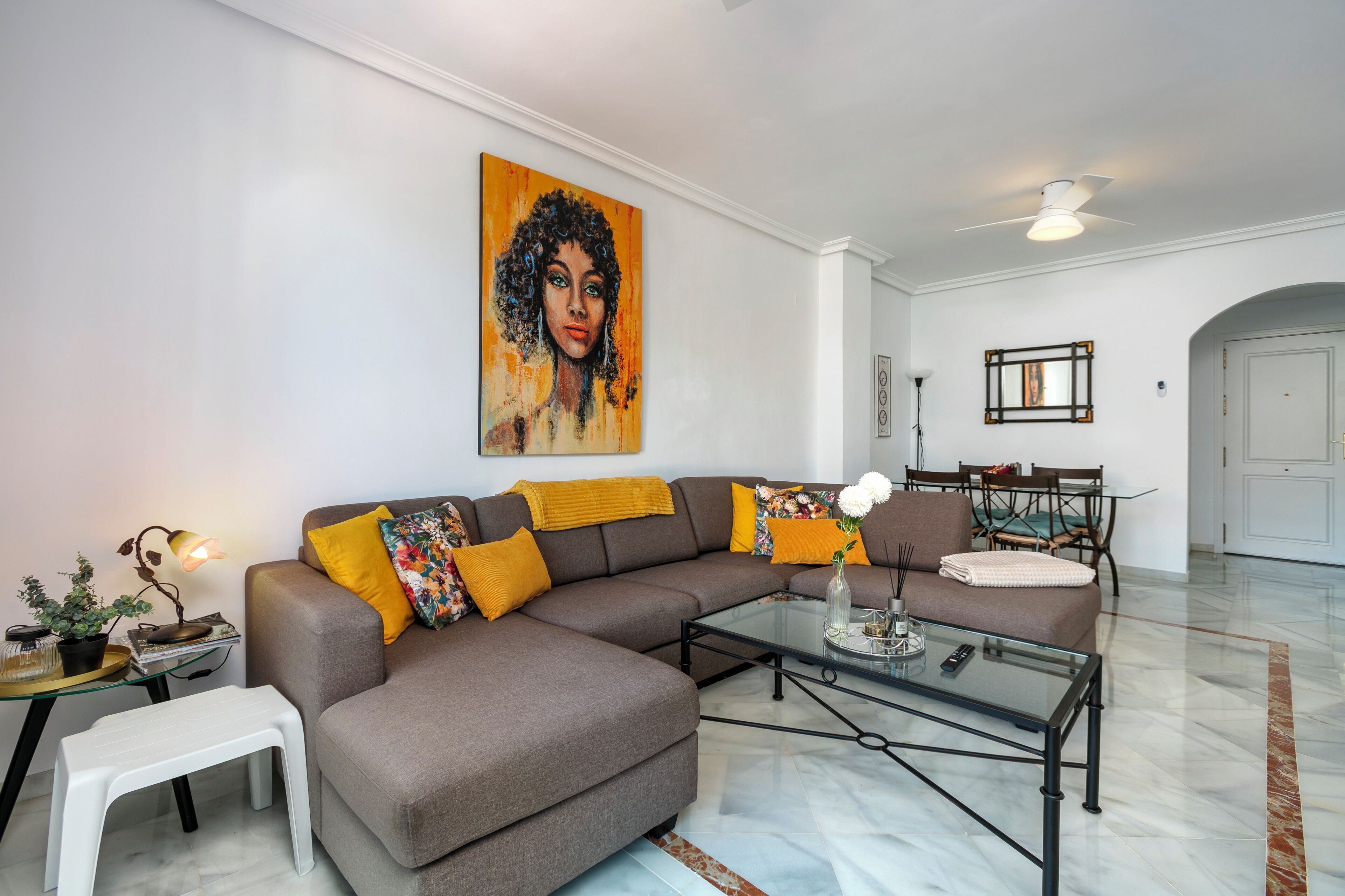 A modern living room with a sectional sofa, a glass coffee table, a dining area with a table and chairs, and a large painting on the wall.