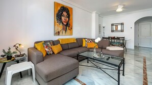 Una sala de estar moderna con un sofá modular, una mesa de centro de vidrio, una zona de comedor con mesa y sillas, y un cuadro grande en la pared.