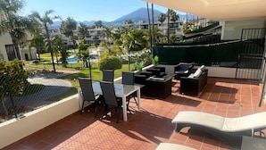 Un patio con una mesa y sillas, sillones de descanso y vistas a una zona turística con palmeras y una montaña al fondo.