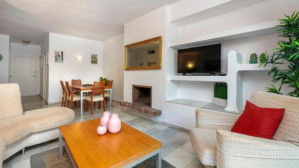 A modern living room with a sofa, a coffee table, a TV mounted on the wall, a fireplace, and a dining area with chairs and a table. - RDR264 - Lovely family Apt close to Puerto Banús (Marbella)