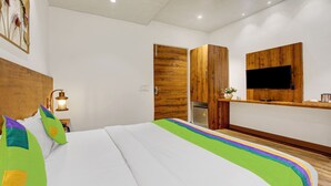 Une chambre d’hôtel moderne avec une tête de lit en bois, un lit agrémenté d’oreillers colorés, une table de chevet avec une lampe, une armoire en bois, un téléviseur fixé au mur et une petite étagère avec un vase de fleurs.