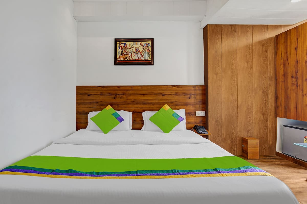 Une chambre d’hôtel avec une tête de lit en bois, un lit recouvert de draps blancs et agrémenté d’oreillers colorés, une petite table de chevet avec un téléphone et un tableau accroché au mur.