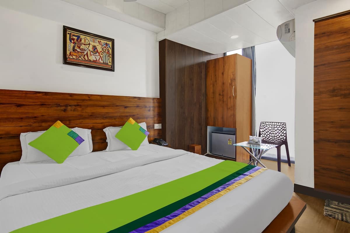 Une chambre d’hôtel avec une tête de lit en bois, un lit avec une literie blanche et des oreillers colorés, une petite table avec une chaise et un climatiseur fixé au mur.