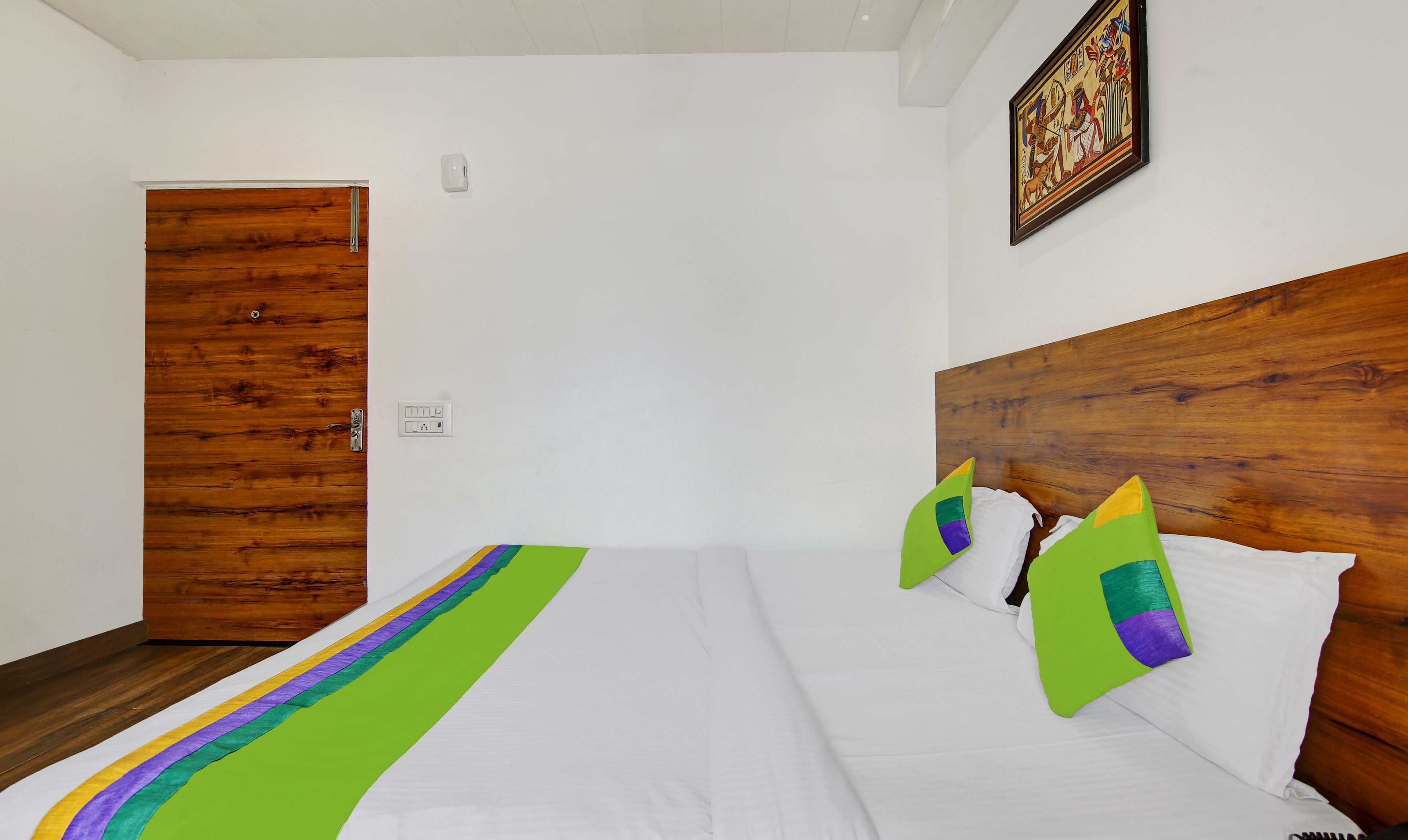 Une chambre d’hôtel avec une tête de lit en bois, du linge de lit blanc et un chemin de table rayé aux couleurs vives.