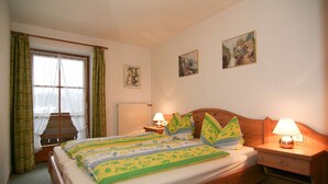 A bedroom with a wooden bed, green and yellow bedding, two bedside lamps, a window with curtains, and two wall-mounted paintings. - Obersee. Apartments Klausner (Schönau am Königssee)