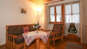 A cozy living room with a wooden ceiling, a sofa, a chair, a table with a floral tablecloth, and a window with curtains. - Obersee. Apartments Klausner (Schönau am Königssee)