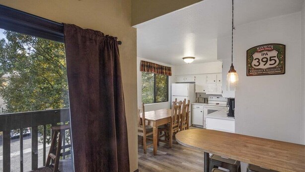 A dining area with a wooden table and chairs, a kitchen with white cabinets, and a large window with a view of trees.