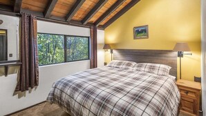 A bedroom with a wooden ceiling, a large window with curtains, a bed with a plaid bedspread, and a nightstand with a lamp.