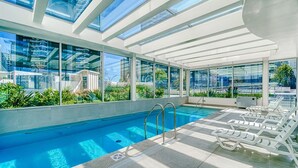 Un espace piscine intérieur moderne, doté d’un plafond en verre transparent, d’une piscine et de chaises longues.