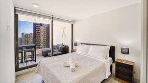 A modern hotel room with a large bed, a small desk, and a balcony with a city view. - Galerna Home 2BR W/Balcony, Wifi & Parking. (Concón)