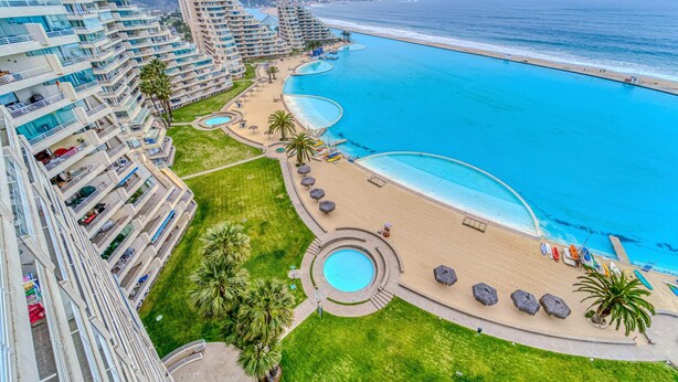 A high-rise hotel with multiple balconies, a central swimming pool, and a beach area with sun loungers. - Bay Dream 2B W/Wifi, Grill & View. (Algarrobo)
