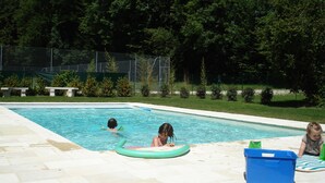 Une piscine où deux enfants jouent, entourée d’une zone délimitée par une clôture, avec des arbres et des bancs.