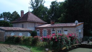 Une maison à deux étages, avec un toit recouvert de tuiles, un petit étang et, au premier plan, des fleurs roses en pleine floraison.