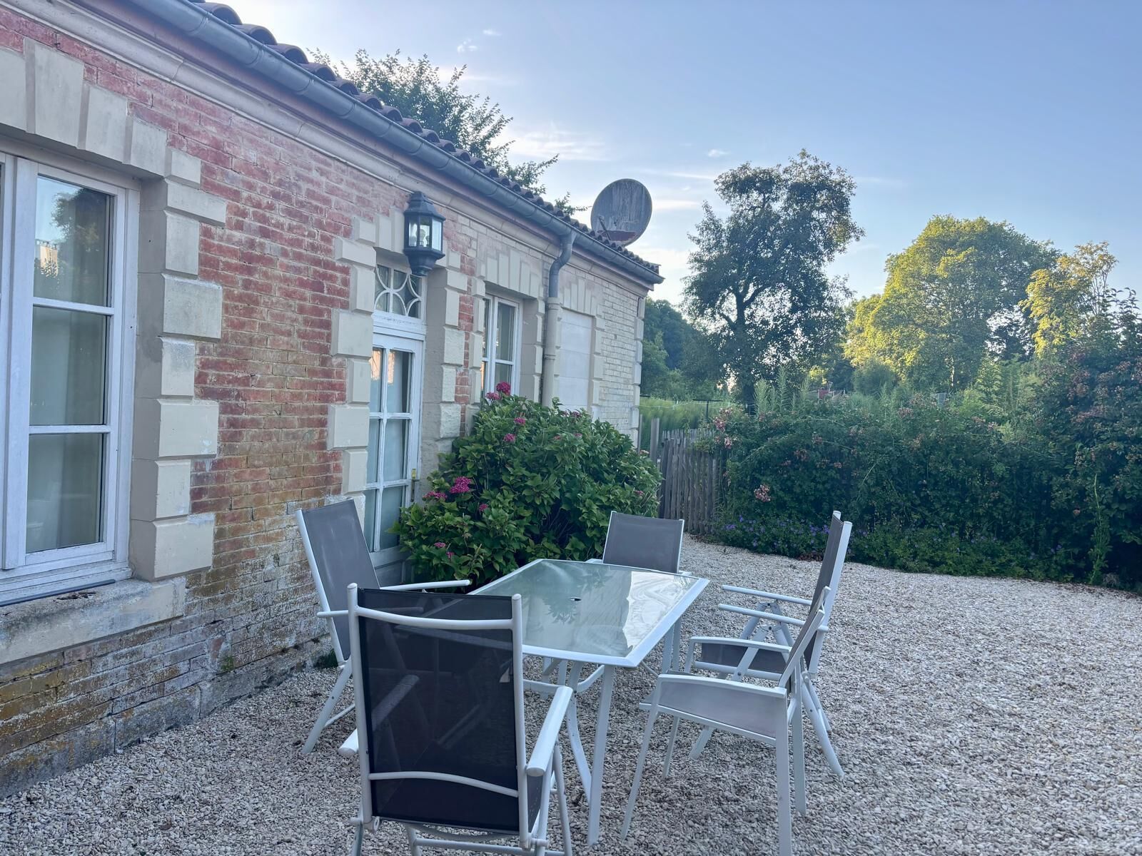 Eine Terrasse mit Tisch und Stühlen, ein Backsteinhaus mit Satellitenschüssel und ein Garten mit Büschen und Blumen.