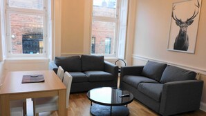 A modern living room with a wooden table, two grey sofas, a round coffee table, and a large window with a view of a brick building.