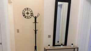 A hallway with a wooden floor, a coat rack, a wall clock, a mirror, and a console table with small plants.