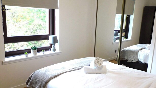 A bedroom with a bed, a window with a view, a bedside table with a lamp, and a mirror. - Charming  2 bed apt close to City and SEC/Hydro (Glasgow)