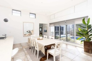 A modern dining area with a large table, white chairs, and a view of a swimming pool through large windows.