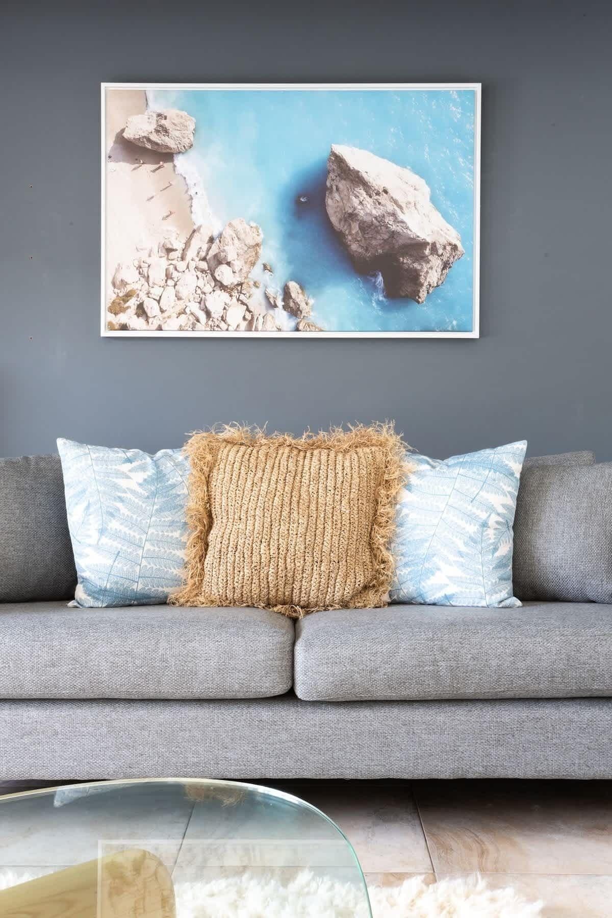 Ein graues Sofa mit einem strukturierten Zierkissen und zwei gemusterten Kissen vor einer Wand mit einem gerahmten Bild.