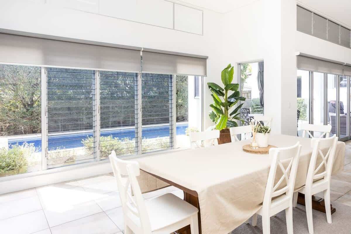 A dining area with a table set for four, large windows with blinds, and a view of a blue body of water.
