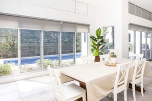 A dining area with a table set for four, large windows with blinds, and a view of a blue body of water. - Magnolia Beach House (Casuarina)