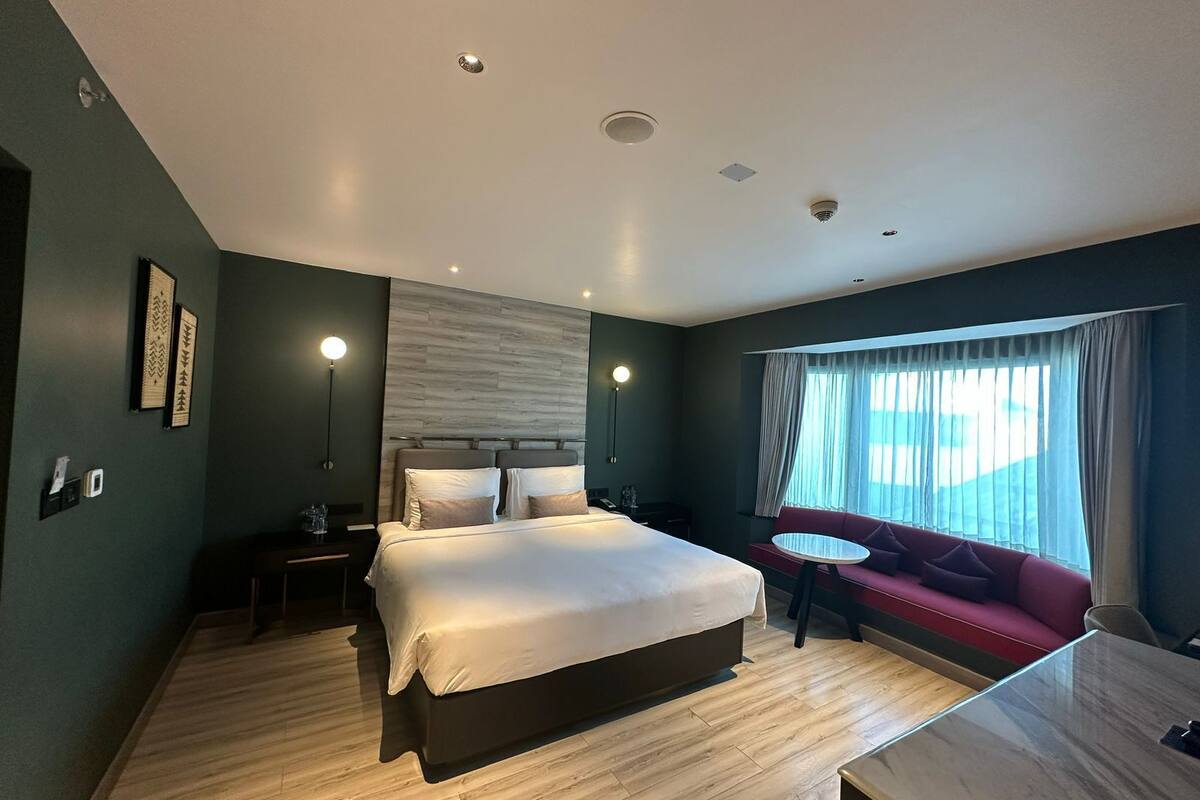Ein modernes Hotelzimmer mit einem großen Bett, einer kleinen Couch, einem Schreibtisch und einem fensterbepflanzten Fenster.