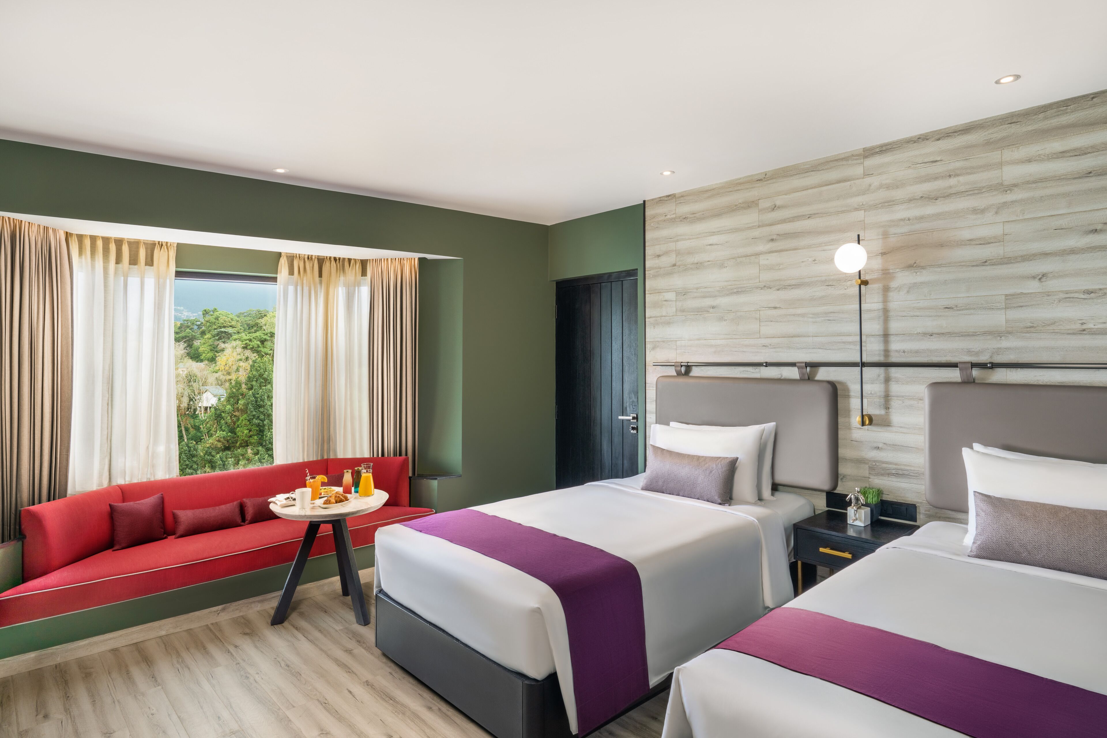 A hotel room with two beds, a sofa, a small table, and a view of greenery.