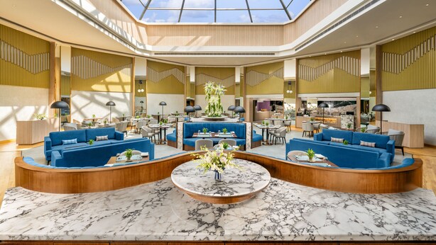 Een ruime lobby met een hoge plafond en dakraam, met blauwe banken, een centrale marmeren tafel en een eetgedeelte op de achtergrond.