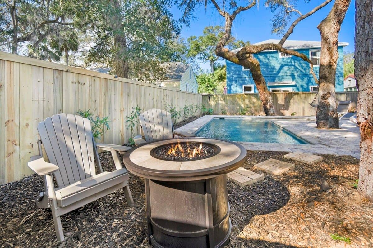 A backyard with a fire pit, two Adirondack chairs, a pool, and a wooden fence.