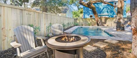 A backyard with a fire pit, two Adirondack chairs, a pool, and a wooden fence.