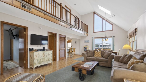A spacious living room with a high ceiling, a wooden balcony, a large TV, and a cozy seating area with a sofa and chairs.