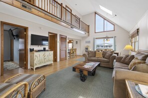Een ruime woonkamer met een hoge plafond, een houten balkon, een grote televisie en een gezellige zithoek met een bank en stoelen.