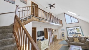 A spacious living area with a wooden staircase, a ceiling fan, a dining area with a table and chairs, and a large window offering a view.