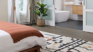 A modern bedroom with a bed, a bedside lamp, a large window with curtains, a potted plant, and a bathroom with a bathtub, shelves, and a mirror.