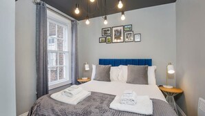 A neatly made bed with a blue headboard, two bedside tables with lamps, and a window with curtains. - Bateman, modern City centre apartment sleeps 6 (York)